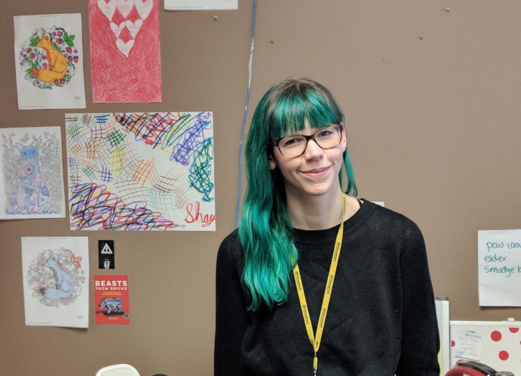 03-march-15-bryn A young white woman with glasses and green hair smiles at the camera.