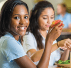 Young woman holding a food item, smiling at the camera. There are more people in the background which is out of focus.