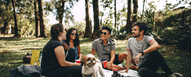 A group of friends sitting on a picnic blanket outside, with a dog.