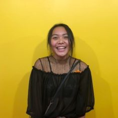 A young woman,standing in front of a yellow wall and smiling.